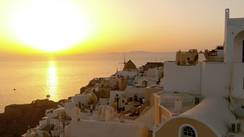 Aerial view of Oia with white buildings and a windmill overlooking the Aegean Sea during a golden sunset in Oia, Greece.