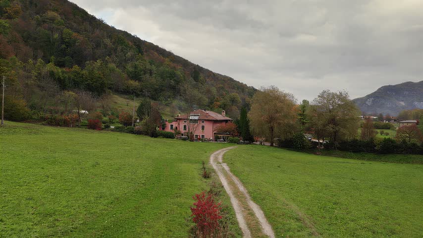 A dirt path winds through a grassy meadow leading toward a traditional house situated at the base of a forested mountain under a cloudy overcast sky