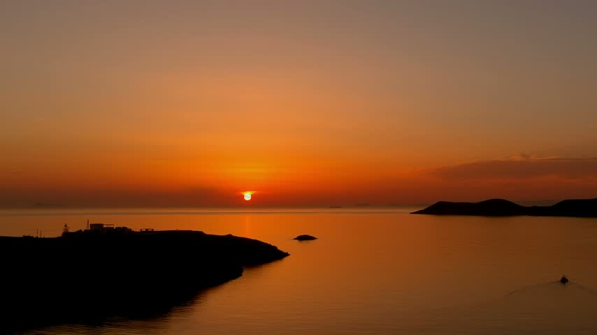Aerial view of a vibrant sunset over the calm sea with the silhouette of a rocky coastline and a small boat in the water near the Islands, Greece.