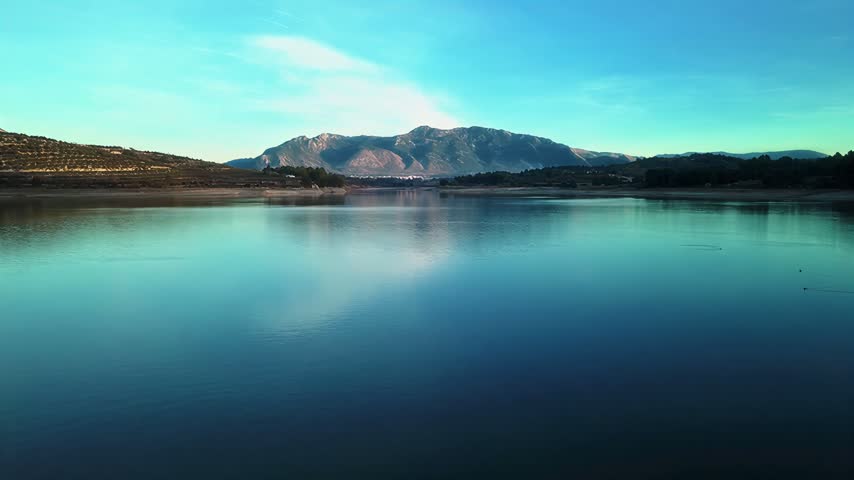 A tranquil landscape featuring calm water mirroring the distant mountain range under a bright sky during the peaceful late afternoon light