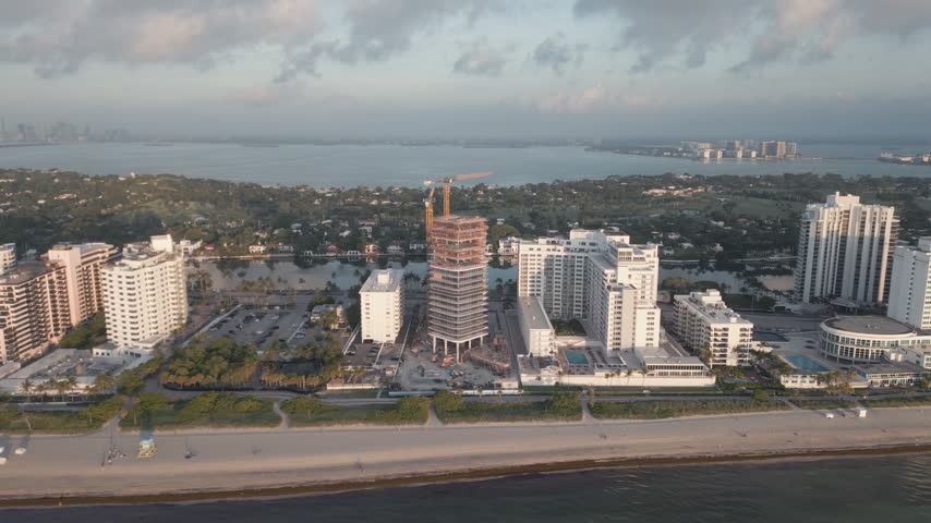 Aerial view of a high-rise building under construction with a crane, surrounded by hotels and residential buildings along the sandy coastline of Miami Beach, Florida, United States.