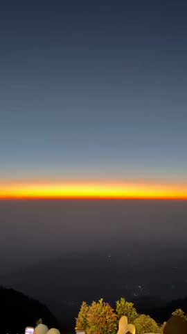 A stunning view of the twilight horizon seen from a mountain peak above the clouds. The vibrant gradient of orange and golden light cuts through the deep blue sky as visitors relax and enjoy the scenic mountain landscape at dawn.