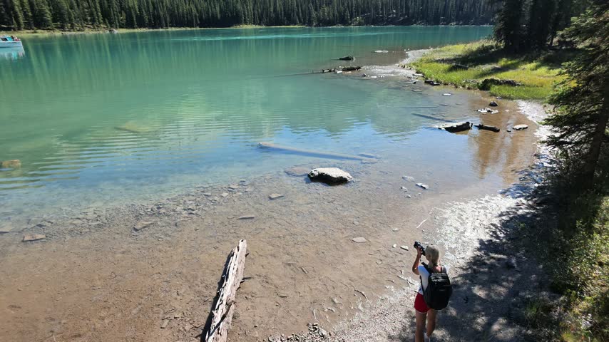Woman standing on shore of Spirit Island in Maligne Lake, holding camera and capturing Spirit Island with its clear water reflecting surrounding forest in Jasper National Park in Alberta of Canada