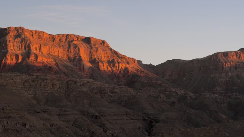 Sunlight slowly disappearing from rugged rocks canyon peaks during serene sunset, illuminating grand rocky landscape with warm and golden glow against a clear twilight sky