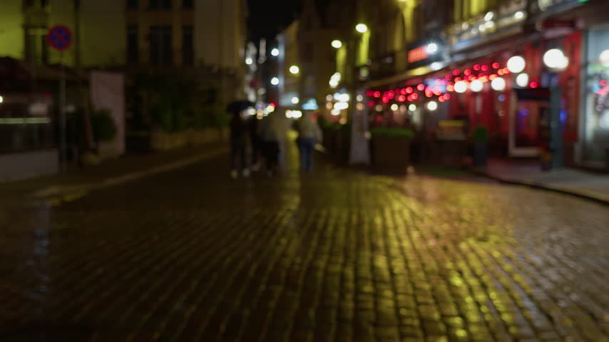 Blurred night street with glowing storefront lights and wet cobblestone reflections in city center