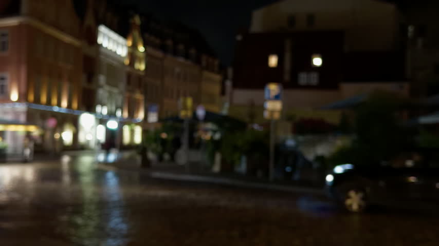 Blurred night street scene with wet pavement, glowing storefront lights, parked car and soft city reflections