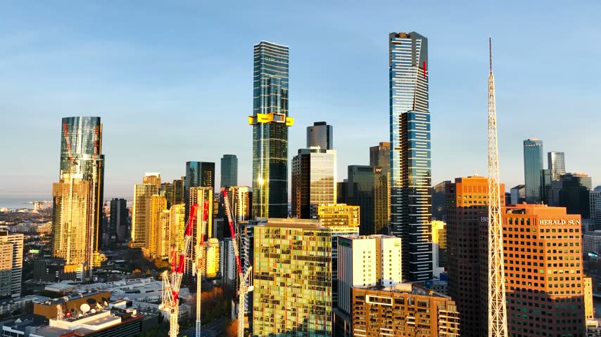 Aerial view of the Melbourne skyline featuring modern skyscrapers, construction cranes, and golden hour sunlight reflecting on glass facades in Melbourne, Victoria, Australia.