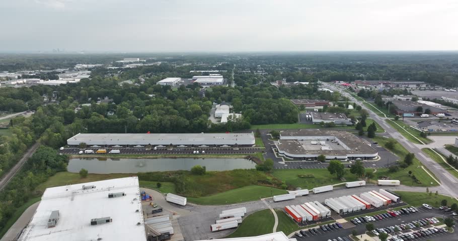 Aerial view of industrial warehouses with parked semi-trucks, a small pond, and surrounding greenery in Indianapolis, Indiana, United States.