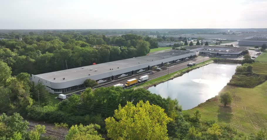 Aerial view of an industrial warehouse facility with a parking lot, trucks, and a pond surrounded by trees in Indianapolis, Indiana, United States.