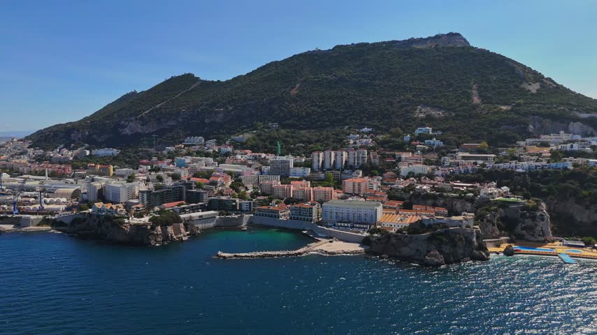 Aerial view of Rosia Bay with its historic coastal buildings, rocky cliffs, and the prominent Rock of Gibraltar under a clear blue sky in Gibraltar, Gibraltar.