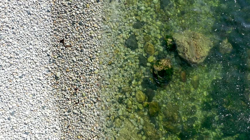 Aerial view of Glenlough Bay featuring a pristine pebble beach meeting the clear green waters of the Atlantic Ocean with submerged rocks in Ardara, County Donegal, Ireland.