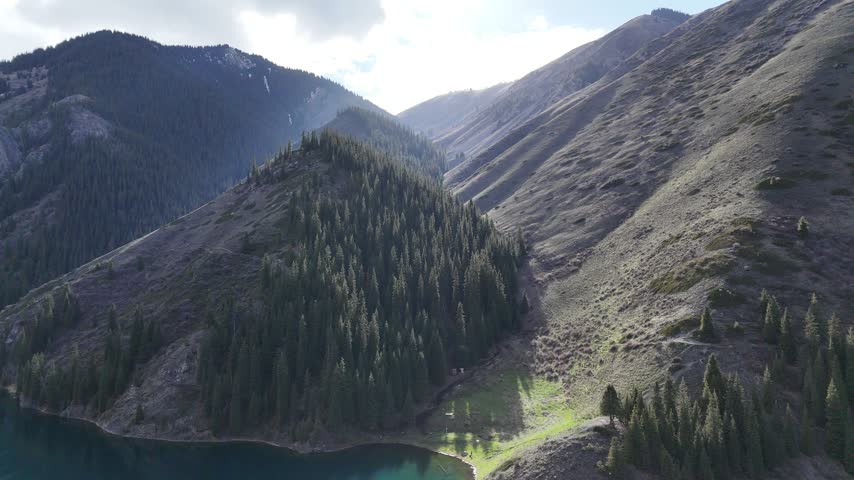Drone view of a peaceful evergreen forest surrounding a turquoise mountain lake in a rugged valley.