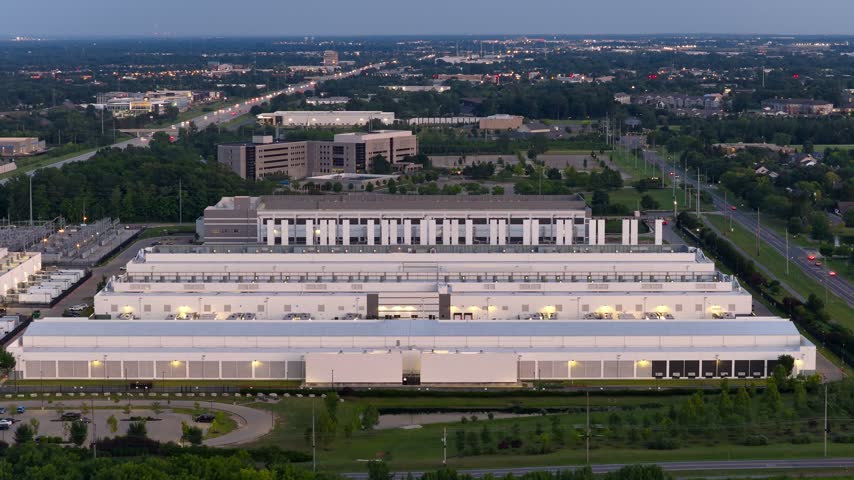 Massive data center campus with surrounding cityscape and highway traffic lights turning on at sunset.