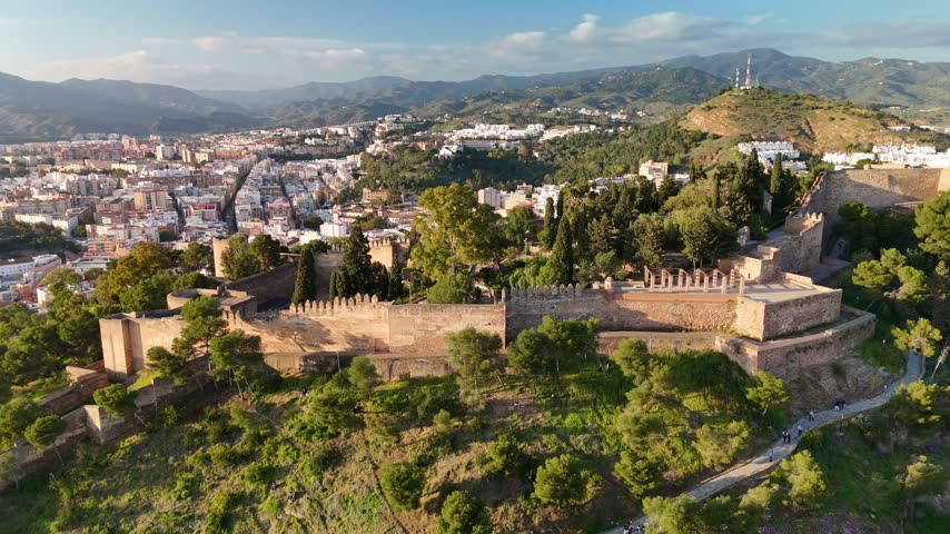 Aerial view of Gibralfaro Castle historic fortress in Malaga, Spain, showing fortress ramparts, defensive wall, wooded hillside, and malaga cityscape. Slide trucking shot