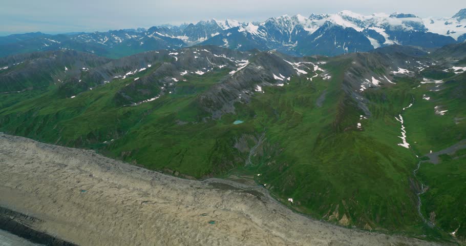 Aerial view of the snow-capped peaks of Mount McKinley and the expansive glacier flowing through the green mountain landscape of Denali Borough, Alaska, United States.