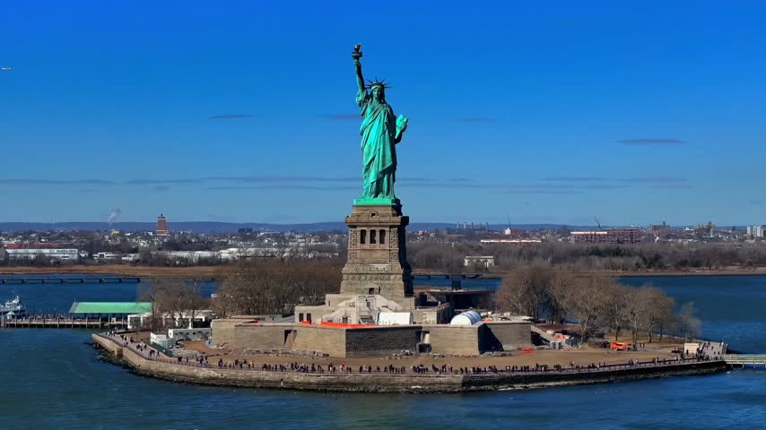 Aerial view of the Statue of Liberty standing on Liberty Island surrounded by the waters of New York Harbor, with the New York City skyline visible in the background under a clear blue sky. Ideal for travel, tourism, landmarks, American culture, history, and cityscape footage.