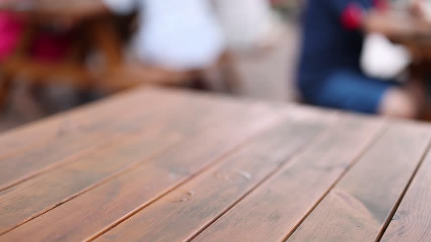Close up shot of male hands placing two chilled pints of craft lager on a rustic wooden table in a busy beer garden setting, cinematic slow motion footage.