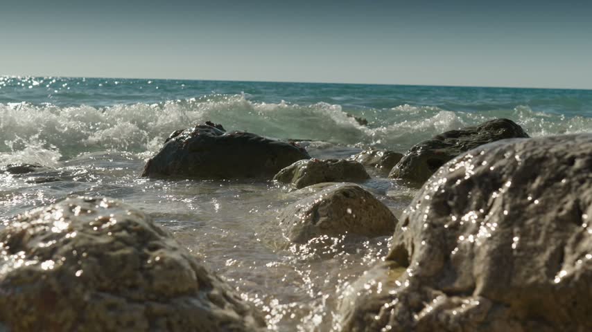 Low angle shot of clear sea waves crashing with white foam over wet coastal boulders in Lefkada Greece.