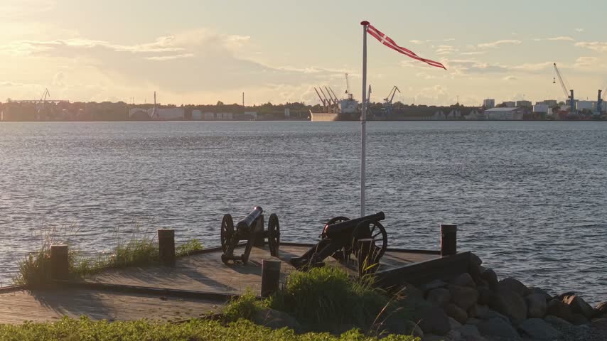 Drone orbit capturing two preserved cast-iron cannons mounted on traditional wooden carriages at the Treårskrigen memorial, overlooking the Little Belt toward Fredericia, Denmark.