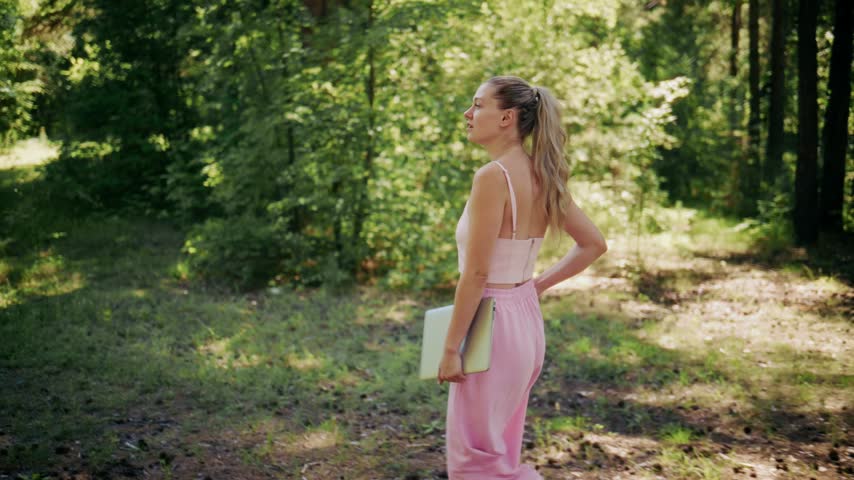 Young woman walks through sunlit forest wearing pink outfit. She holds books, smiles softly, and looks relaxed. Gentle sunlight filters through green trees. Ideal for education, nature