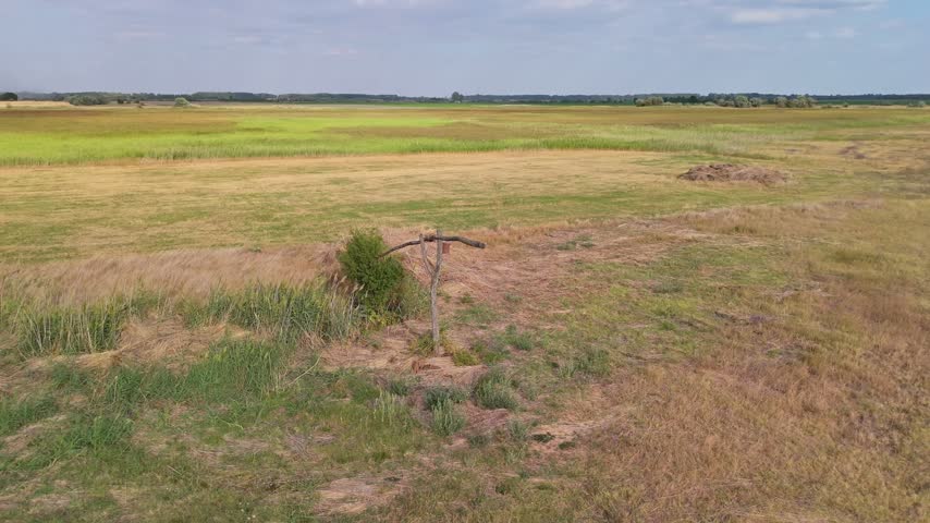 A weathered wooden post stands amid dry grasses and green meadowland in the flat rural landscape near Újsolt, Hungary, with scattered trees beneath a lightly clouded sky.