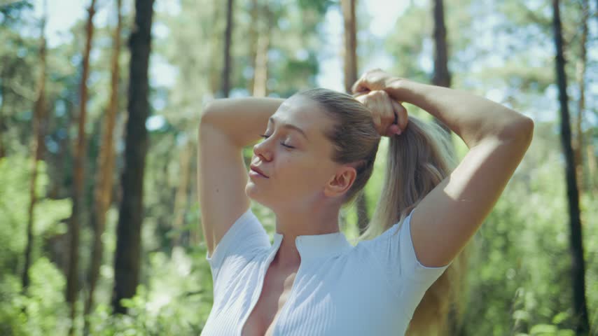 Young woman ties blonde hair in forest. Sunlight filters through tall pine trees. She looks calm and refreshed. Nature surrounds her in peaceful greenery. Ideal for wellness, self-care