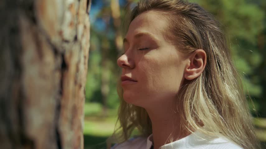 Woman leans against a pine tree trunk, eyes closed. She breathes deeply, calm and peaceful. Sunlight filters softly through surrounding greenery. Her face shows serene contentment