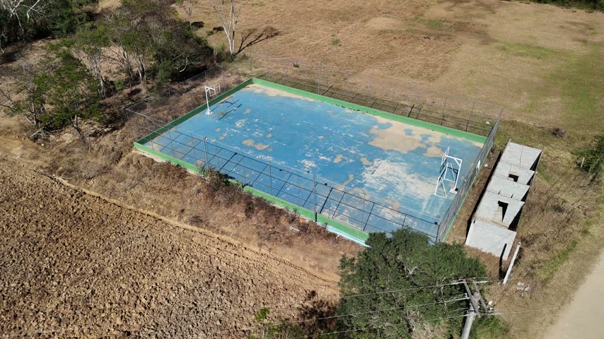 Aerial view of a weathered blue basketball court surrounded by a chain-link fence in a rural landscape with dry fields and trees in Nova Friburgo, Rio de Janeiro, Brazil.