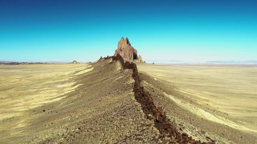 Aerial drone flight following a dramatic volcanic dike toward Shiprock in northwestern New Mexico. The dark rock wall leads across the vast high desert toward the towering Ship Rock volcanic formation under a clear blue sky.