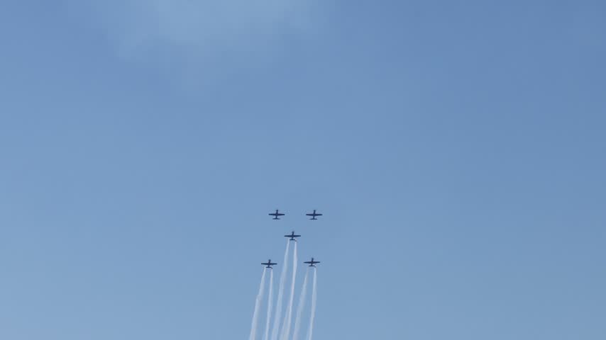 Six propeller aircraft perform an impressive aerial display, flying in formation against a clear blue sky, leaving long white smoke trails, showcasing precision and skill at an airshow.