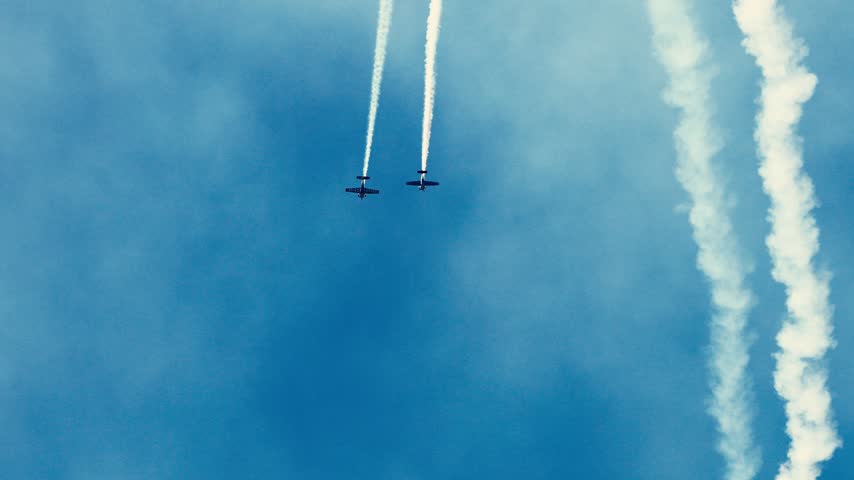 Two planes fly in formation, performing aerobatic stunts and leaving smoke trails. Dynamic aerial display against clear blue sky.