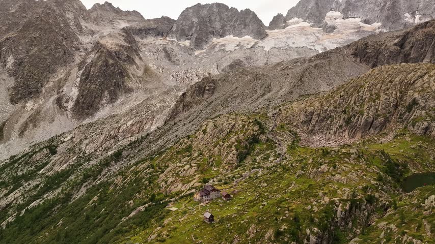 Aerial view of the rugged Presanella mountain range featuring rocky slopes, alpine meadows, and a small mountain hut nestled in the landscape of Giustino, Trentino-Alto AdigeS, Italy.