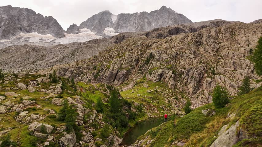 Aerial view of Presanella mountain landscape with rocky slopes, green vegetation, and a person in a red jacket standing by a small alpine lake in Giustino, Trentino-Alto Adige, Italy.