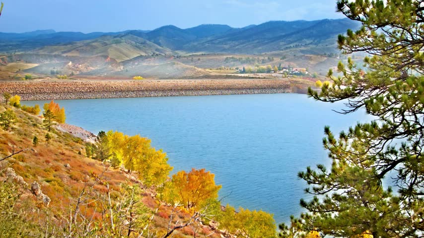 A calm blue reservoir sits surrounded by colorful autumn hills and distant mountain ranges.