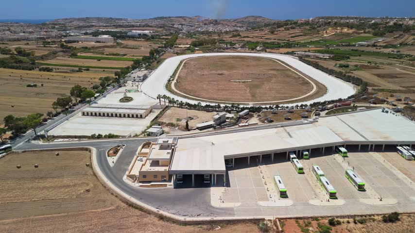 Xewkija, Gozo Park and Ride bus terminal with buses, parking area and surrounding countryside fields in Malta, viewed in a sideways tracking aerial shot over the transport hub.