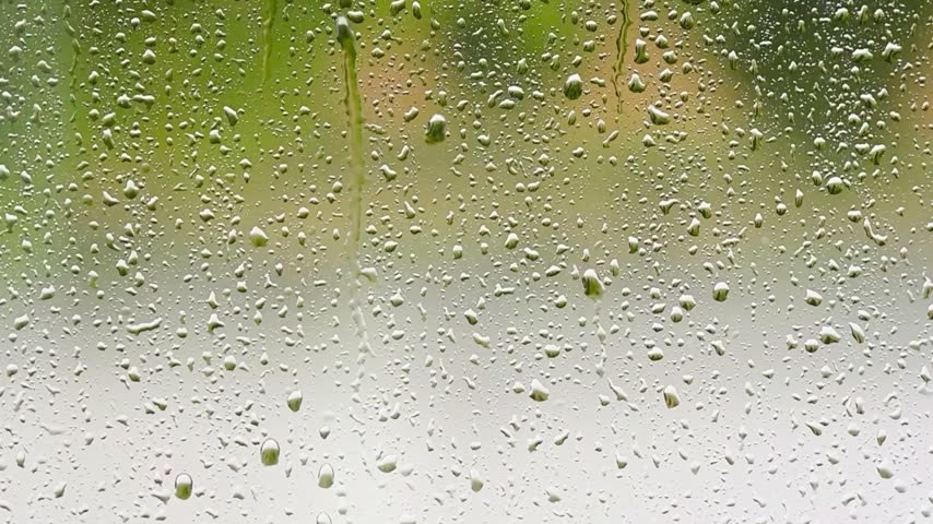 Raindrops accumulate and stream down a transparent glass surface against a blurry green background.