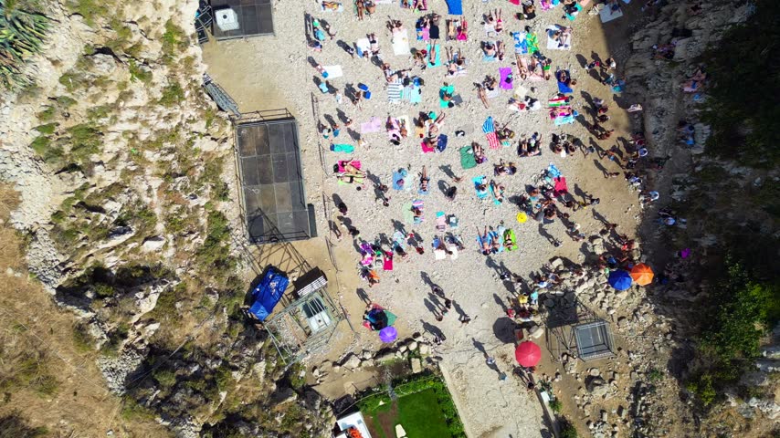 Aerial view of Polignano a Mare beach with people sunbathing on the pebbly shore of Lama Monachile beach surrounded by rocky cliffs in Polignano a Mare, Apulia, Italy.