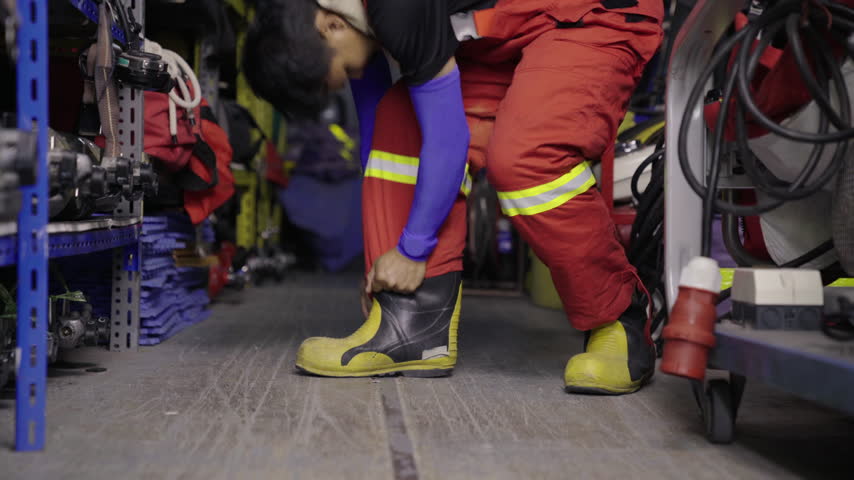Asian rescue worker putting on orange protective jumpsuit, preparing for emergency response surrounded by helmets and rescue gear inside station equipment room.