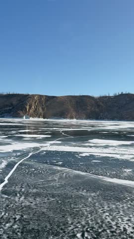 Slow motion vertical video of car driving on ice road of frozen Baikal Lake. View from cabin on ice surface and rocky coast of Olkhon Island on sunny winter cold day. Auto travel and adventure