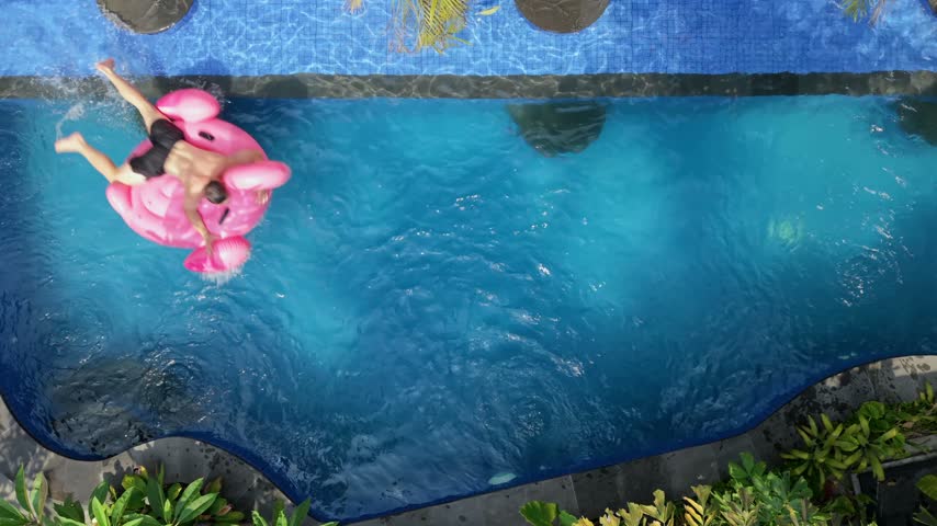 Top-down view of a man jumping onto a pink flamingo float, drifting in a swimming pool and falling into the water. Fun summer vacation, leisure and playful lifestyle concept.