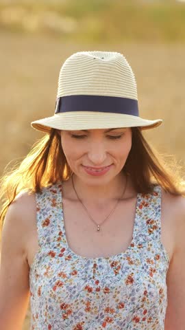 Charming young woman wearing a straw hat and floral dress smiling playfully at the camera while standing in a golden wheat field at sunset, with beautiful backlight highlighting her hair