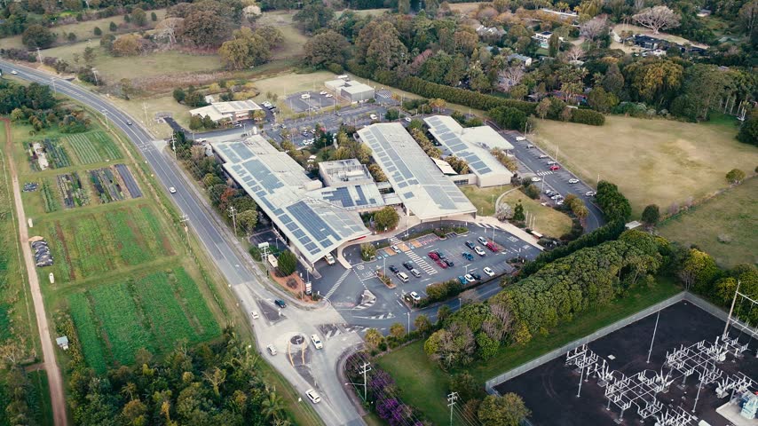 Aerial view of a hospital complex with solar panels, adjacent highway, power substation, and surrounding farmland in Byron Bay, New South Wales, Australia.