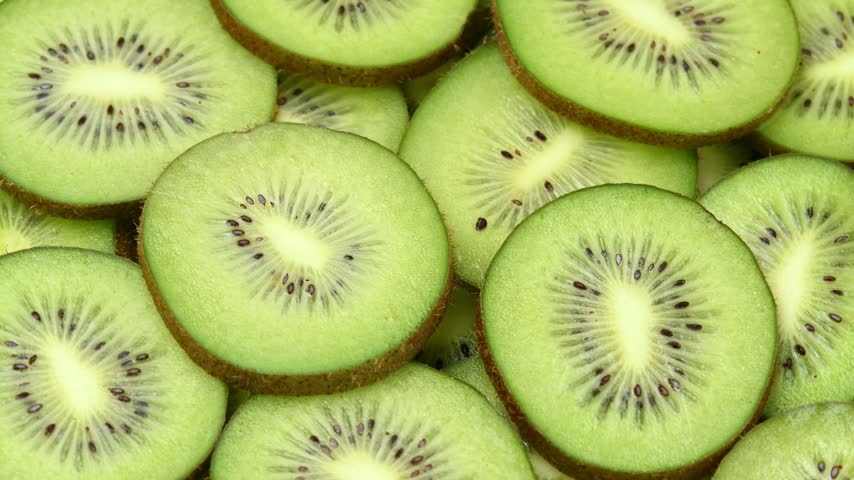 Overhead kiwi slices rotate in a horizontal frame, revealing green flesh, seeds, and fresh texture.