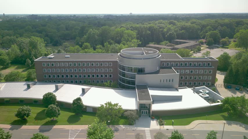 Drone shot orbiting a modern university of Michigan buildings in Dearborn, surrounded by a lush green campus on a sunny day.