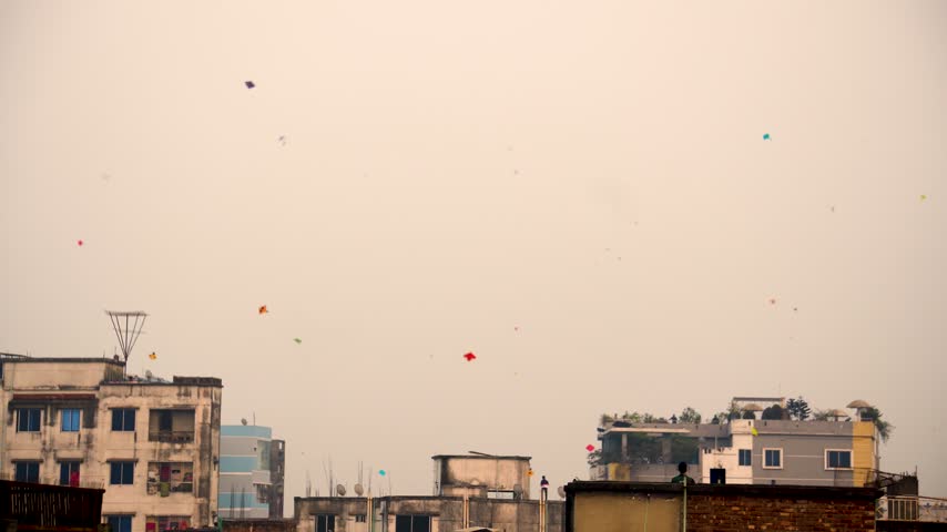 Wide shot of vibrant colorful kites flying across the evening sky above Old Dhaka rooftops, with people gathered on the rooftops during the annual Sakrain kite flying festival in Bangladesh.