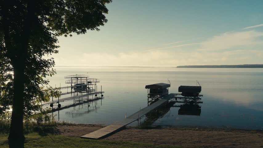 Calm lake in the early morning with docks, boat lifts, a sandy shoreline, and trees reflecting on the still water
