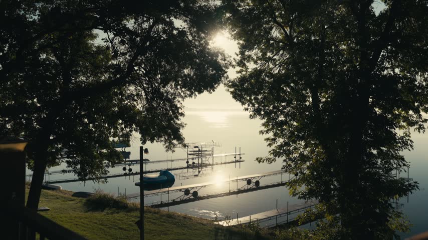 Morning sunlight shines through trees over a calm lake with docks and boat lifts along the shoreline