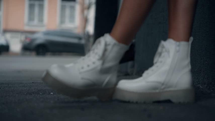 Close up of person wearing trendy white platform boots walking on city sidewalk during bright daylight with blurred urban street background showing car and building exterior in soft focus style
