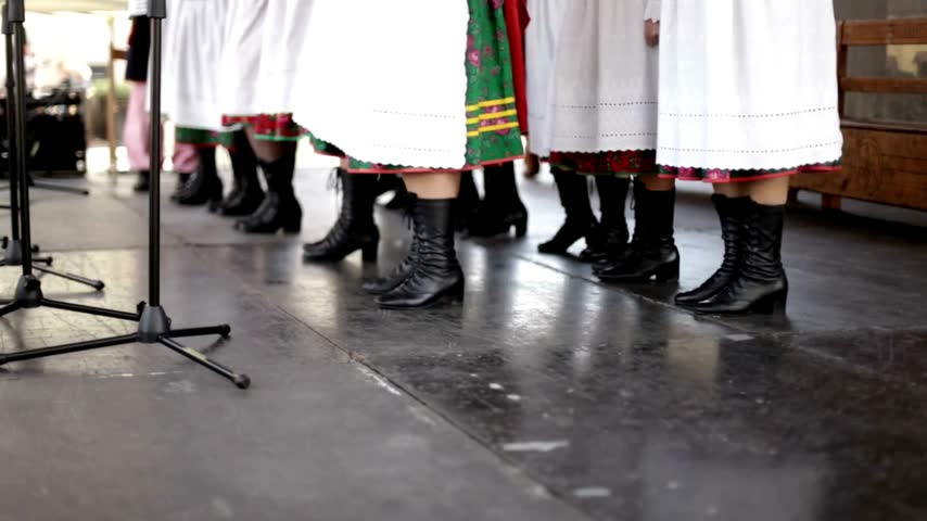 Performers standing in rows wearing traditional white skirts and black leather boots while participating in a cultural dance performance on a wooden outdoor stage during a daytime celebration