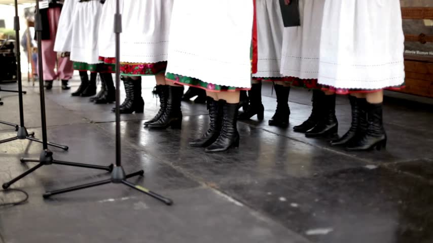 Performers wearing traditional white skirts and black leather boots standing on an outdoor stage during a cultural festival event in Poland as part of a folk music group performance outdoors